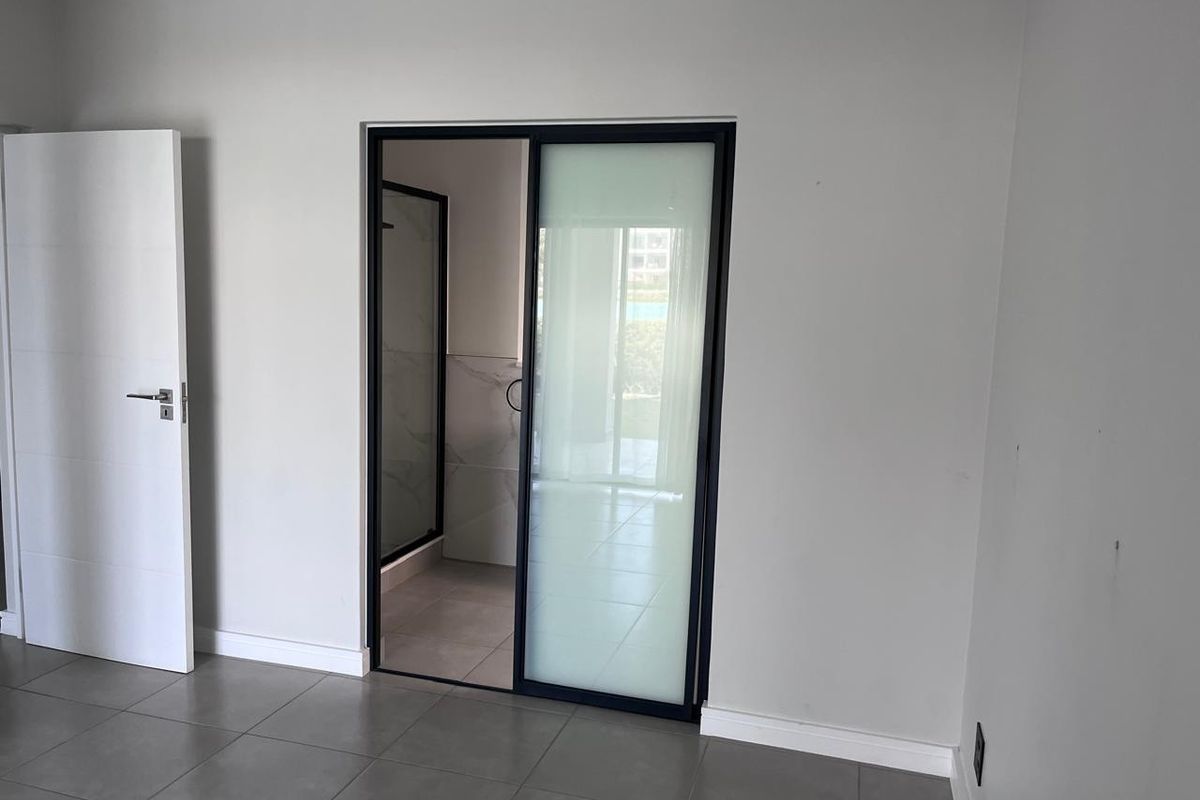 A clean, modern bedroom featuring neutral wall tones, large format floor tiles, and a contemporary sliding glass door leading to an en-suite bathroom.