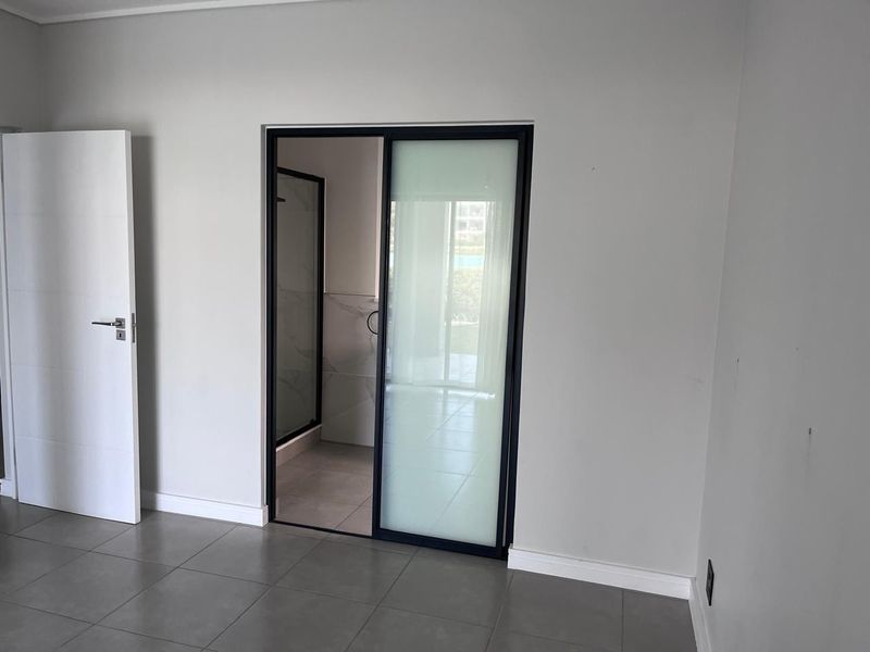 A clean, modern bedroom featuring neutral wall tones, large format floor tiles, and a contemporary sliding glass door leading to an en-suite bathroom.