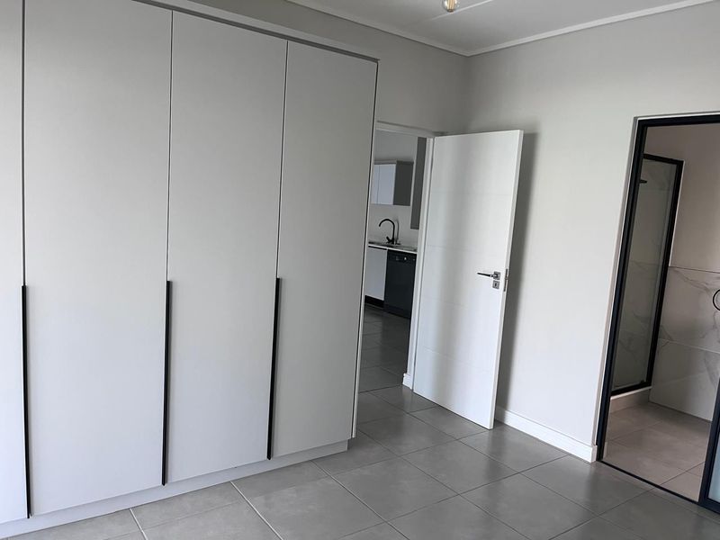 A modern, empty bedroom featuring neutral tiled flooring, built-in cupboards with sleek black handles, and direct access to an adjoining kitchen and bathroom.