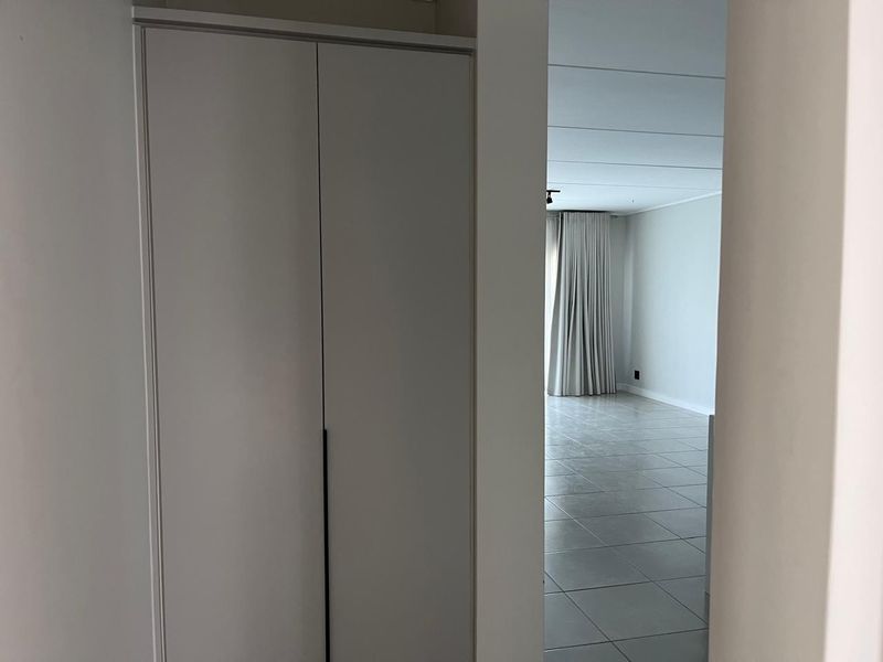 A clean, neutral hallway featuring a built-in cupboard and tiled flooring, leading into an open-plan living area.