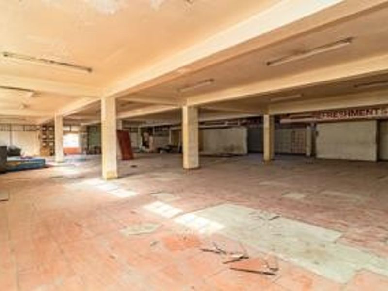 A large, vacant open-plan commercial space featuring concrete columns, tiled flooring, and industrial fluorescent lighting, requiring significant refurbishment and cosmetic updates.