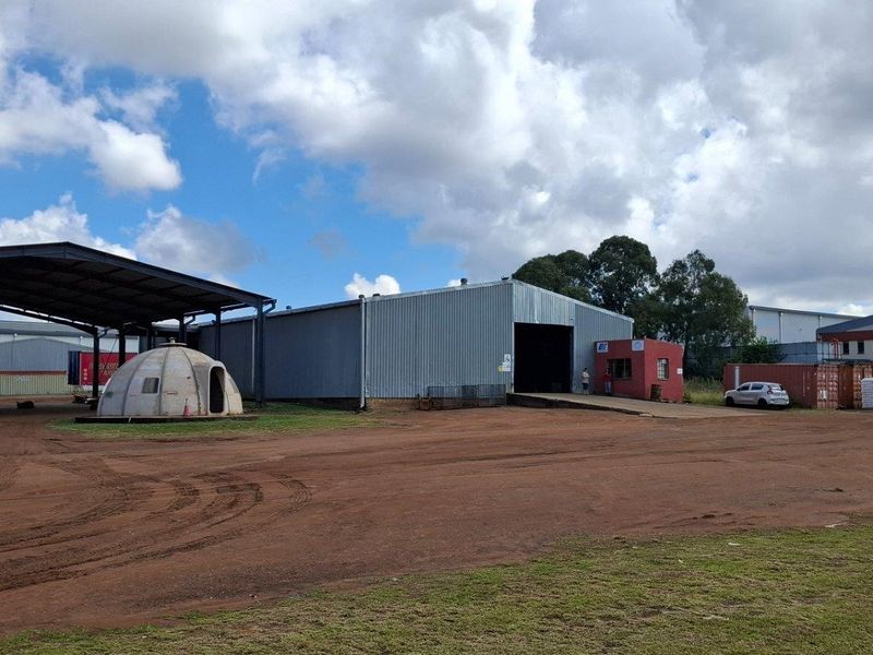 Industrial property featuring a large metal warehouse, covered loading bay, concrete ramp, and security hut situated on an expansive unpaved yard.