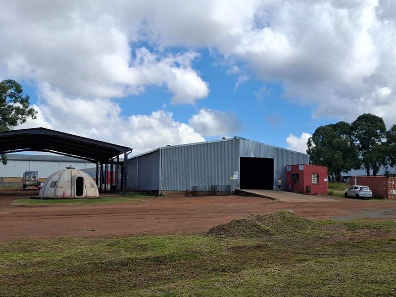 Industrial property featuring a large metal warehouse, an open-sided steel canopy, a loading ramp, and a security guard booth on an unpaved site.