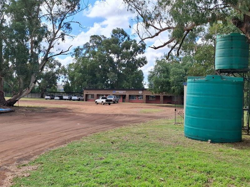 A view of the property frontage featuring a large unpaved driveway, mature trees, and prominent water storage tanks on the site.