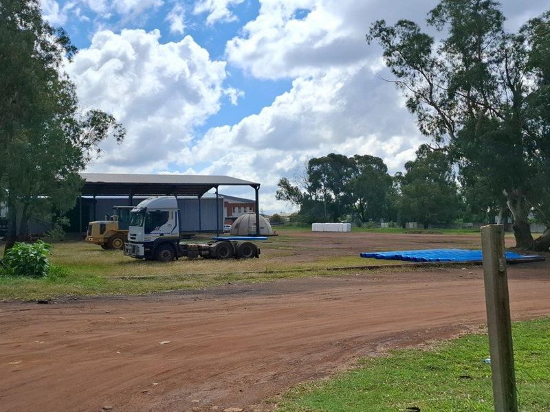 A large, open vacant residential plot featuring an unpaved access road, mature trees, and an open-sided metal storage shed structure.