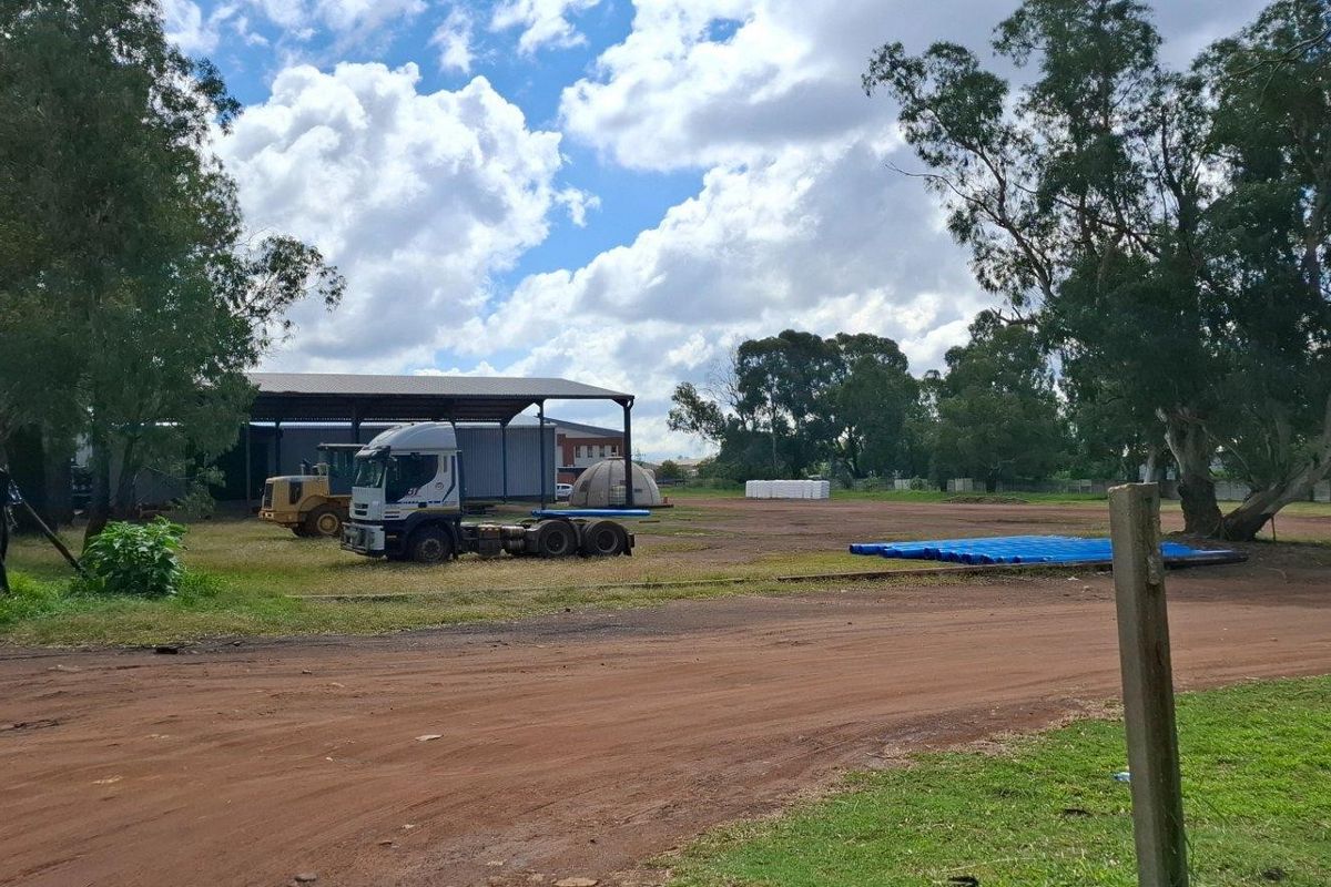 A large, open vacant residential plot featuring an unpaved access road, mature trees, and an open-sided metal storage shed structure.
