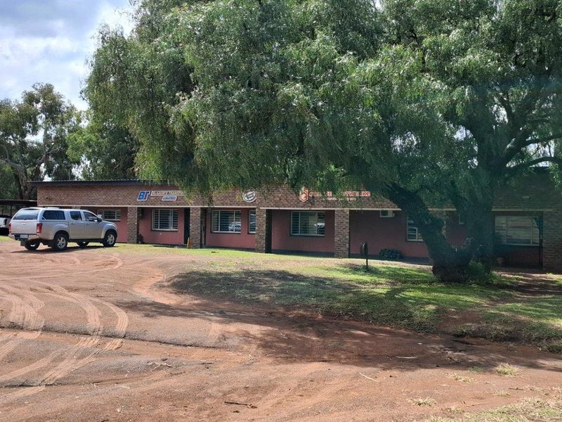 Front view of a single-story brick building with a covered veranda, situated on a large plot with mature trees and an unpaved driveway.