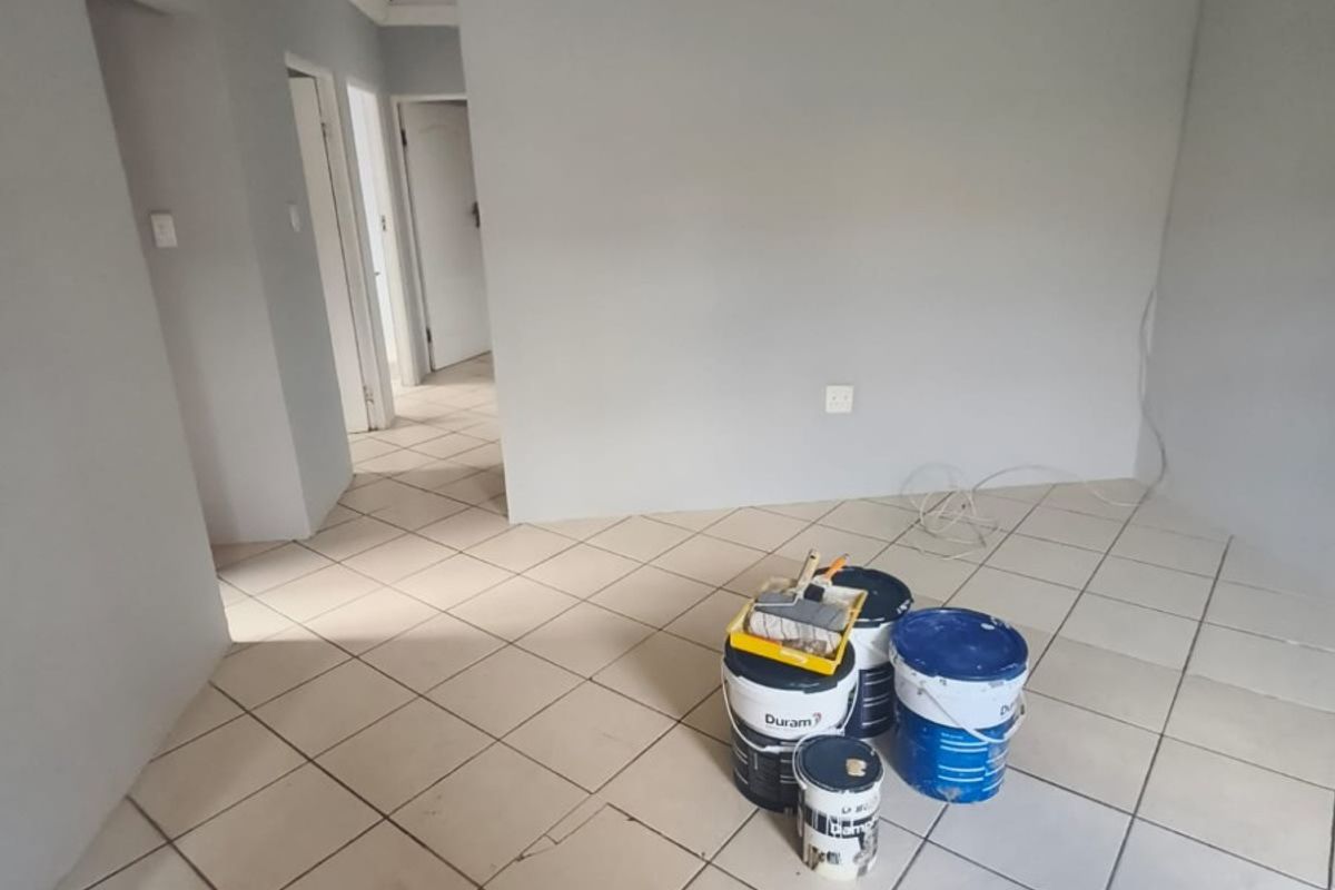 An empty living area featuring neutral grey walls, ceramic floor tiles, and decorative ceiling cornicing, currently undergoing minor maintenance.