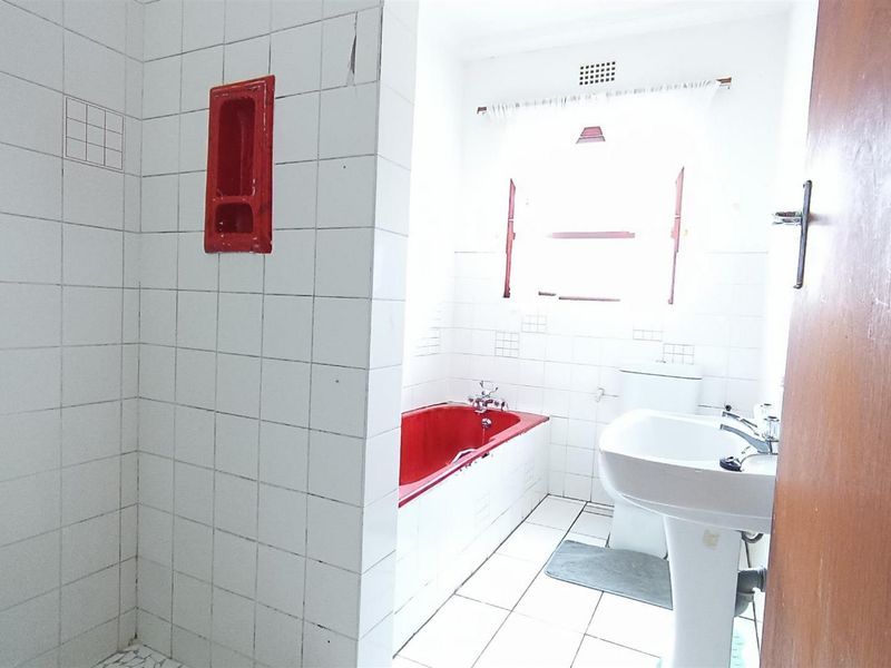 A dated bathroom featuring white wall tiles, a red built-in bathtub, pedestal basin, and toilet, requiring cosmetic updates to modernize the space.