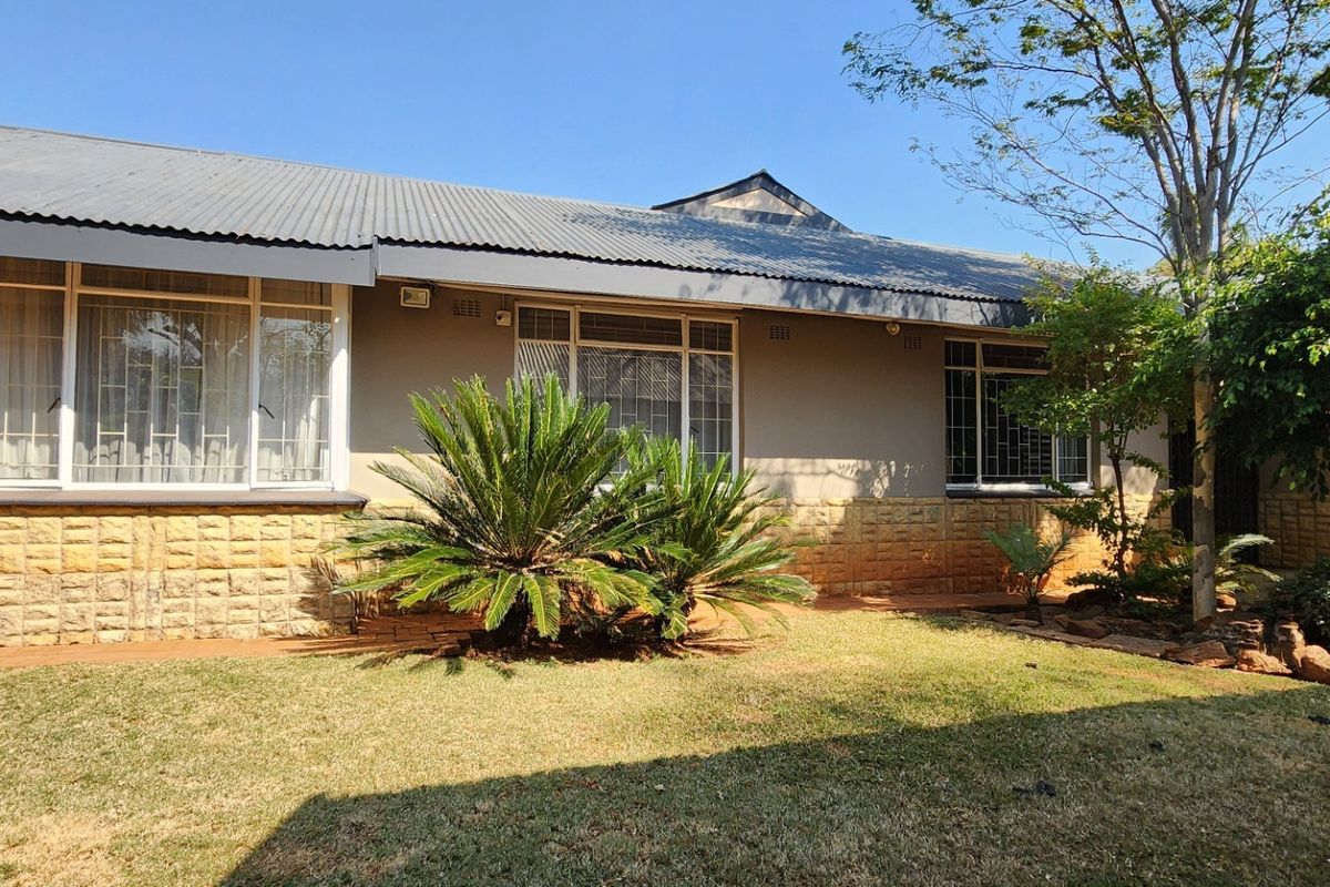 Rear view of the house featuring a corrugated metal roof, stone cladding base, and large windows with security bars overlooking a garden.