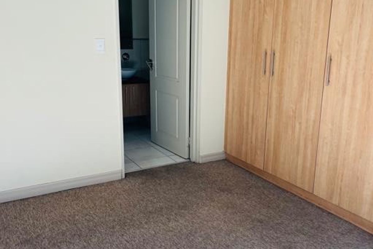 A clean, empty bedroom featuring neutral walls, carpeted flooring, and built-in wooden cupboards, with an adjacent view into a bathroom.