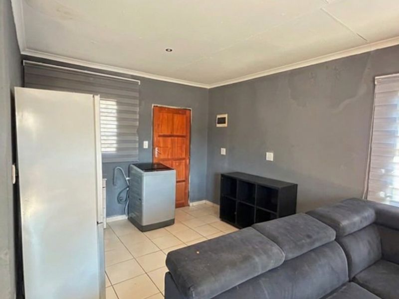 A living area featuring tiled flooring, roller blinds, and a wooden door, showing signs of wall surface wear and water staining.