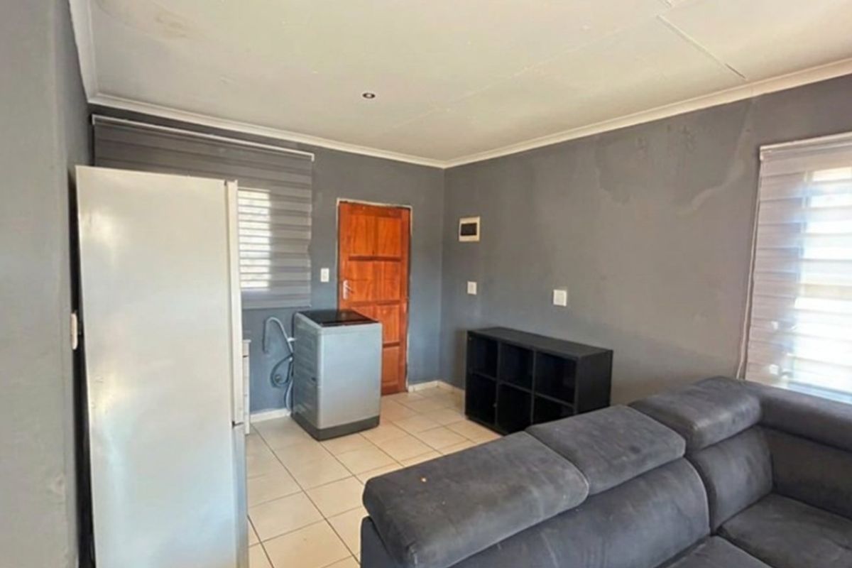 A living area featuring tiled flooring, roller blinds, and a wooden door, showing signs of wall surface wear and water staining.