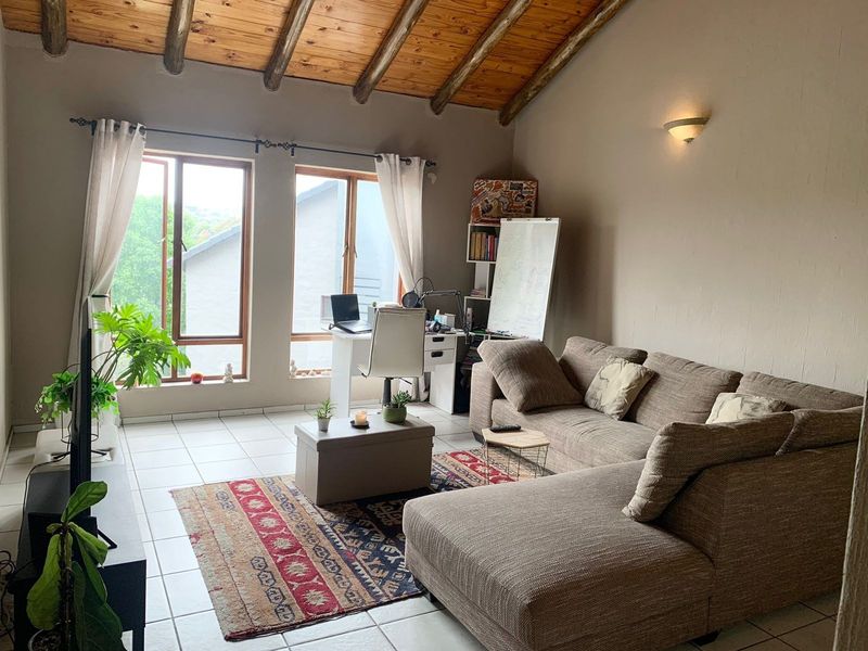 A spacious living room featuring a high vaulted ceiling with exposed timber beams, tiled flooring, and large windows providing natural light.