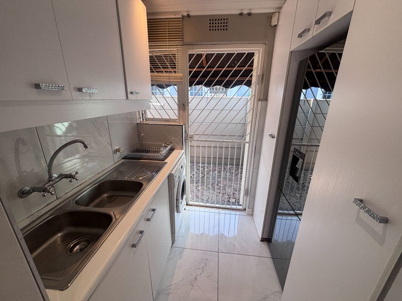 Compact scullery featuring white built-in cabinetry, double stainless steel sink, tiled flooring, and direct access to an outdoor area via a security gate.