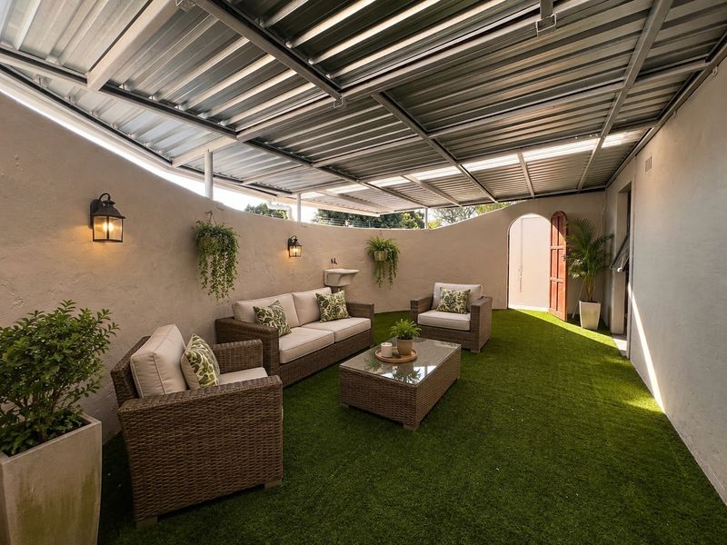 A covered outdoor patio area featuring a curved wall, artificial grass flooring, and a wall-mounted basin under a corrugated metal roof structure.