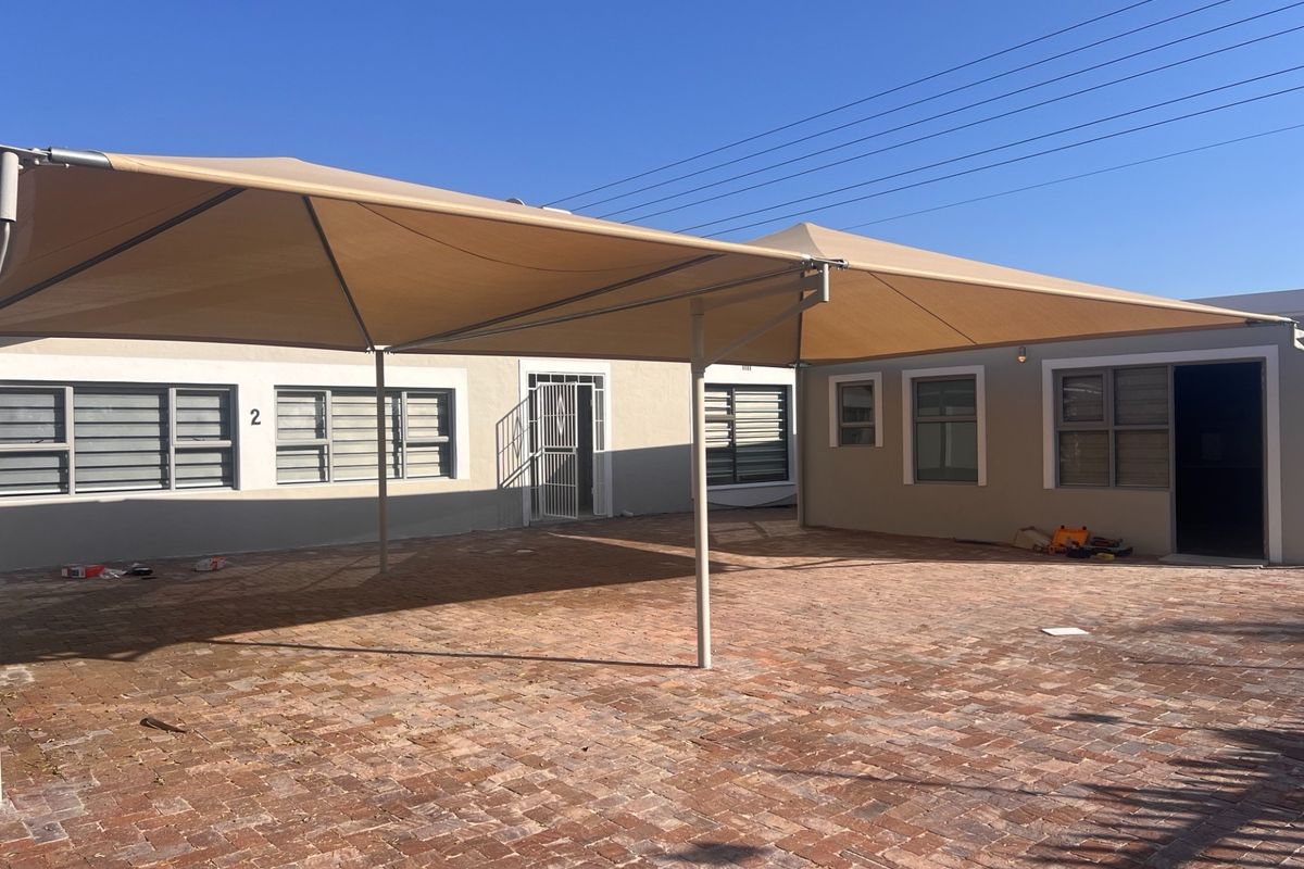 A paved rear courtyard area featuring a large shade sail structure, security gates, and aluminium window frames.