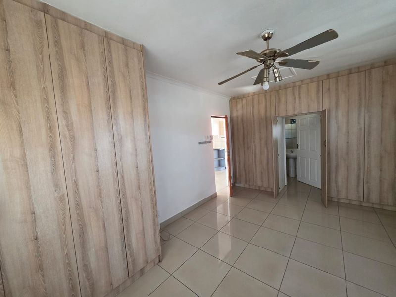 Spacious bedroom featuring extensive built-in wooden cupboards, neutral tiled flooring, and a ceiling fan, offering a clean and functional living space.