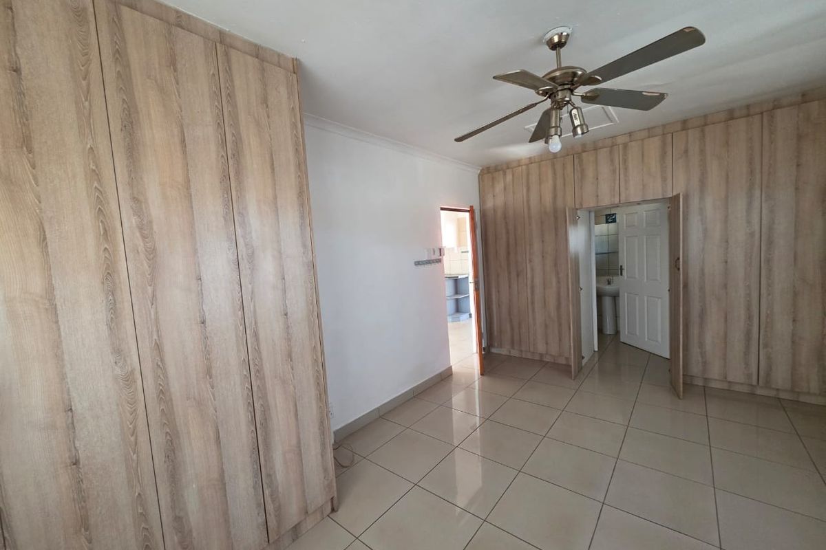 Spacious bedroom featuring extensive built-in wooden cupboards, neutral tiled flooring, and a ceiling fan, offering a clean and functional living space.