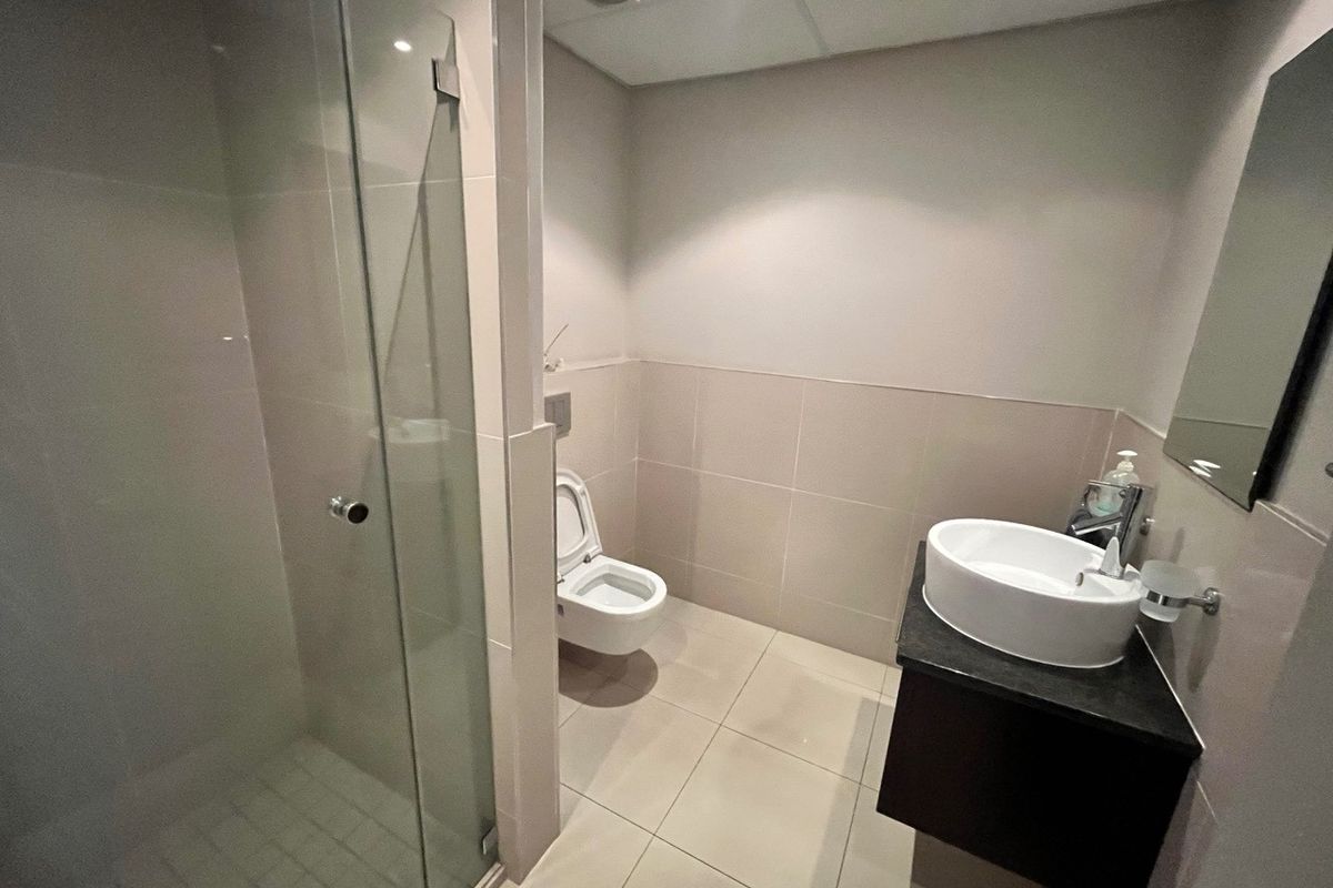 A modern, clean office bathroom featuring a glass-enclosed shower, wall-mounted toilet, and a contemporary vessel sink with a vanity cabinet.
