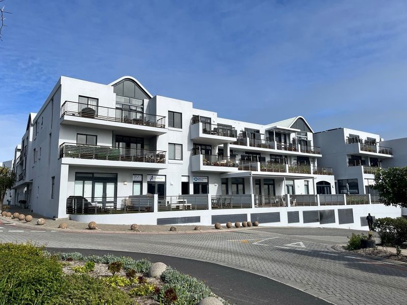 A modern multi-story office building featuring white exterior finishes, multiple balconies with metal railings, and ground-level access with paved driveway frontage.