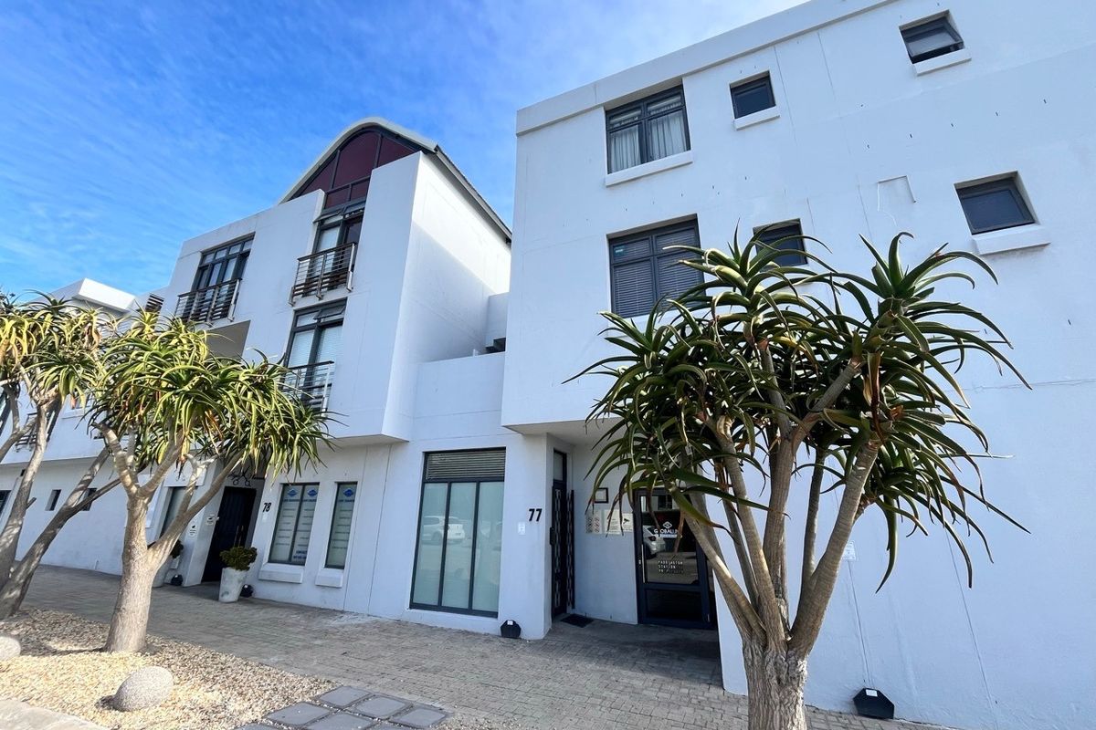 Modern multi-story office building facade featuring white exterior walls, aluminium window frames, and paved entrance area with established landscaping.