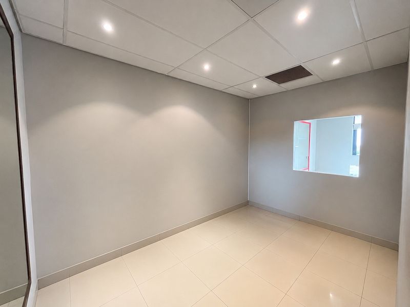 A clean, empty office space featuring neutral walls, tiled flooring, a suspended ceiling with recessed lighting, and an internal window.