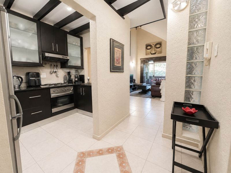 A functional kitchen featuring dark cabinetry, built-in oven and hob, and exposed ceiling beams, flowing into an open-plan living area with glass block accents.