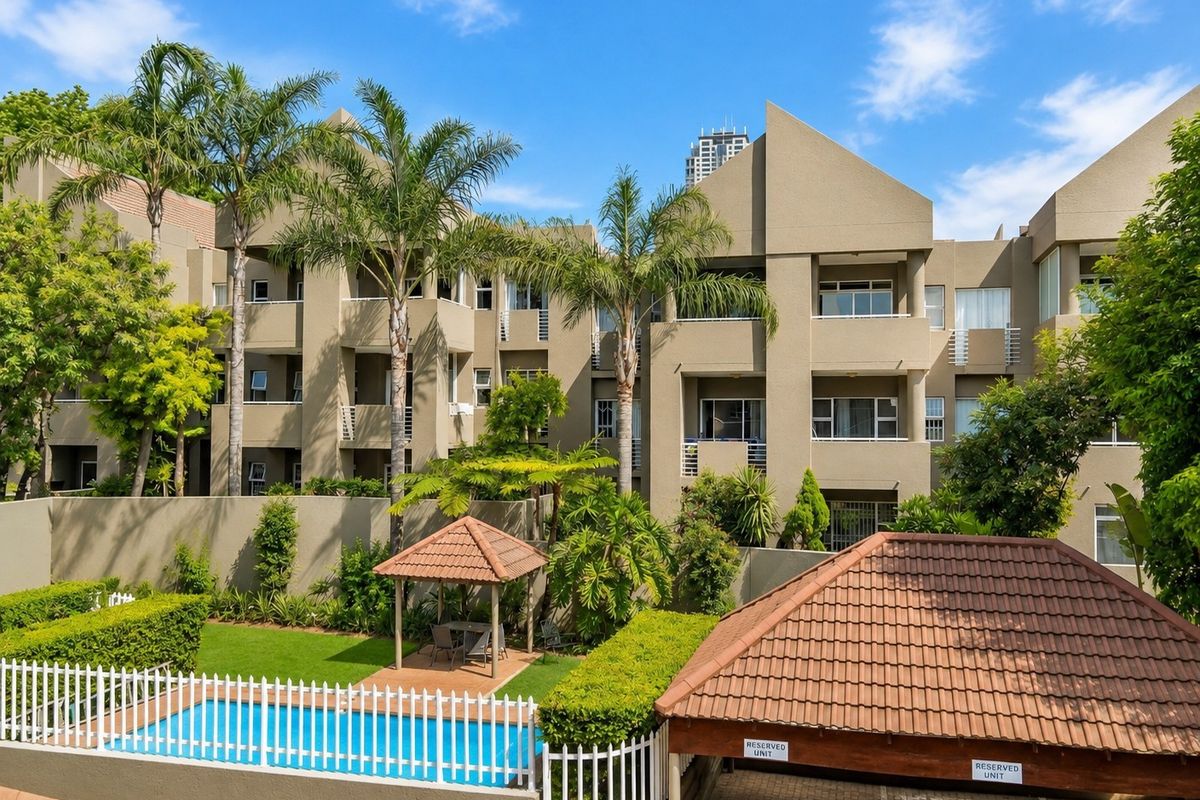 Exterior view of an apartment complex featuring a swimming pool, gazebo, landscaped garden, and covered parking area.