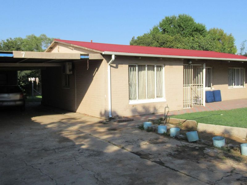 Front view of a single-story brick house featuring a metal roof, attached carport, and concrete driveway with security gate access.