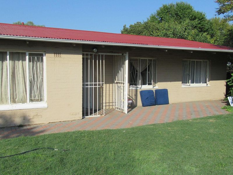 Rear view of a single-story brick house featuring a red corrugated metal roof, security gates, and a paved patio area overlooking the garden.