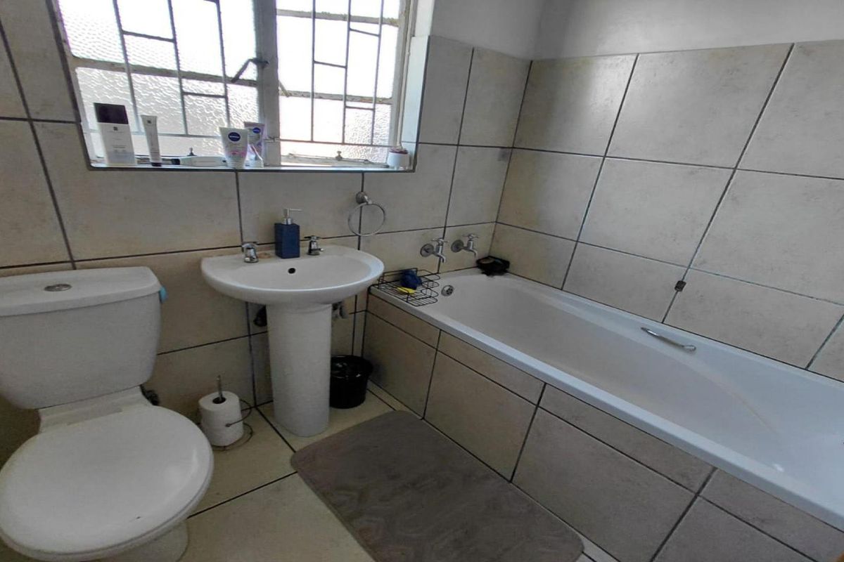 A functional bathroom featuring a pedestal basin, toilet, and bathtub with tiled finishes and a window with security bars.