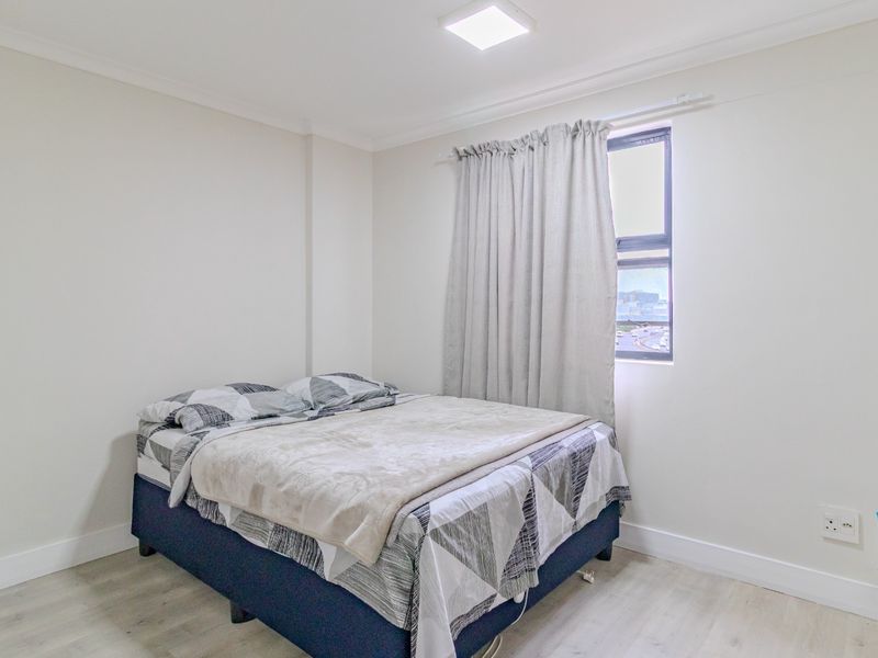 A clean and neutral bedroom featuring light laminate flooring, coving, and a window with natural light.