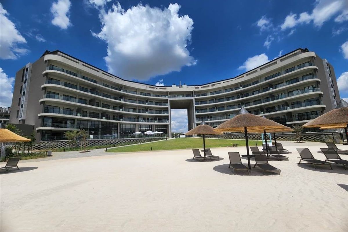 A wide-angle view of the apartment complex rear facade, featuring multiple levels of balconies overlooking a landscaped lawn and sandy beach area.