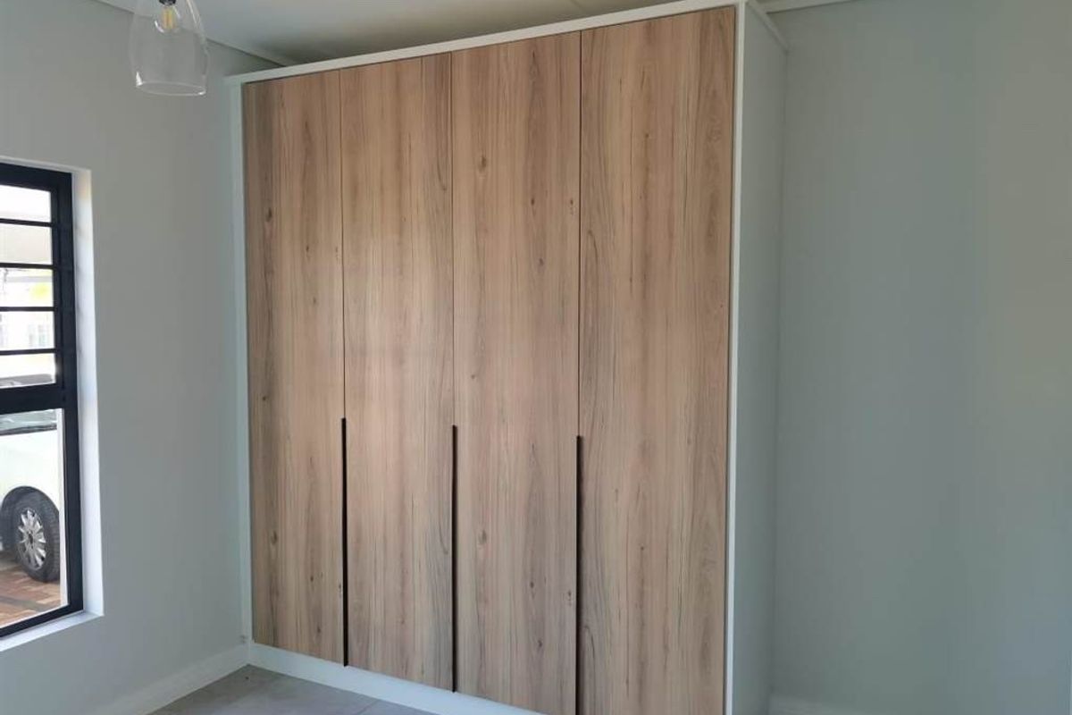 A bright bedroom featuring modern wood-finish built-in wardrobes, neutral tiled flooring, and a contemporary pendant light fixture.