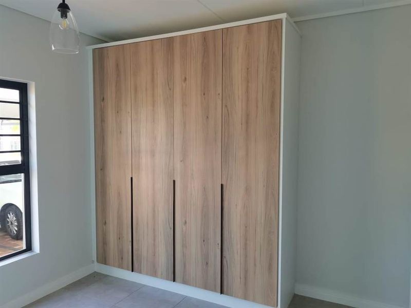 A bright bedroom featuring modern wood-finish built-in wardrobes, neutral tiled flooring, and a contemporary pendant light fixture.