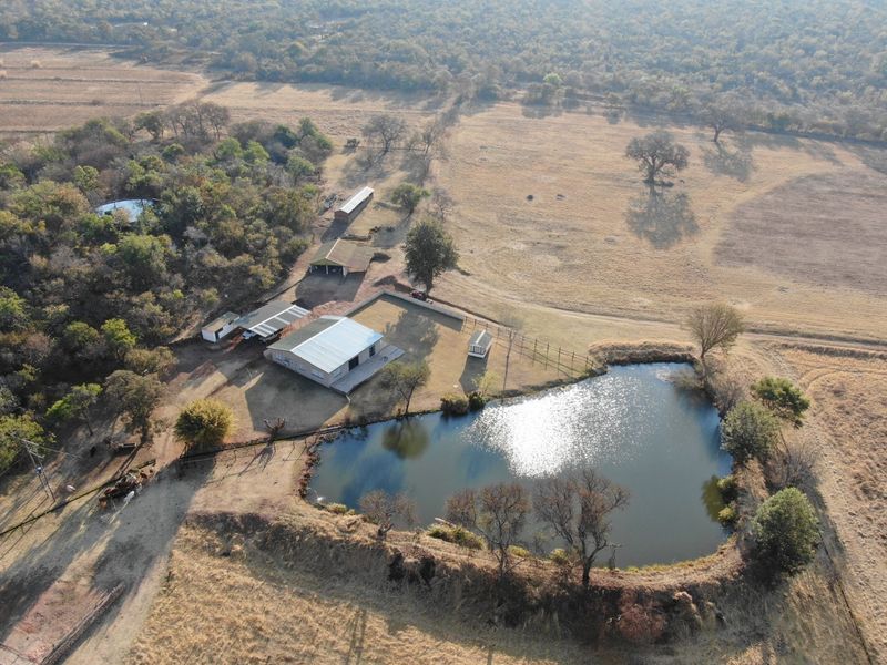 Aerial view of a farm property featuring a large dam, main dwelling with a metal roof, and various outbuildings set within open grazing land.