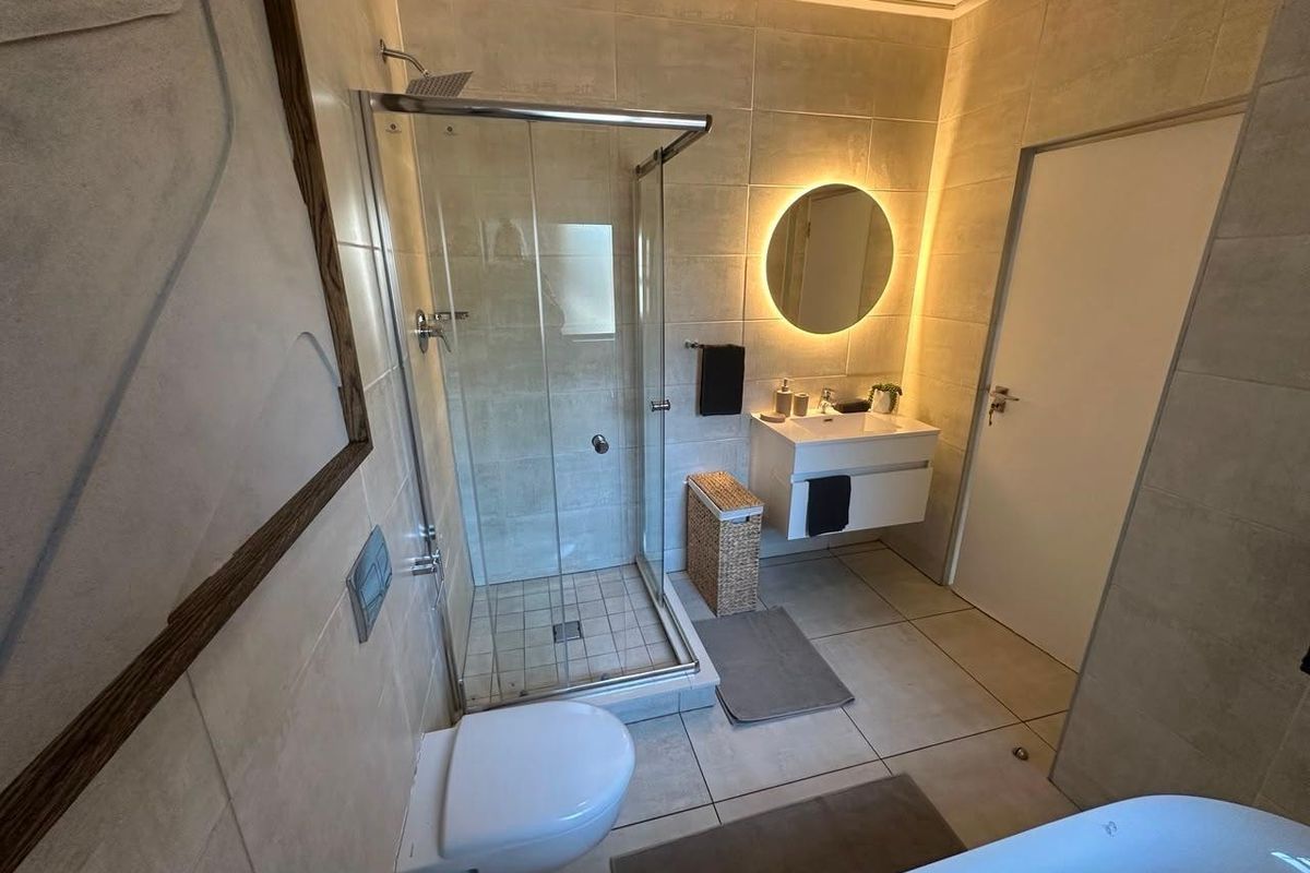 Modern bathroom featuring a floating vanity, backlit circular mirror, wall-mounted toilet, and a glass-enclosed shower with neutral floor-to-ceiling tiling.