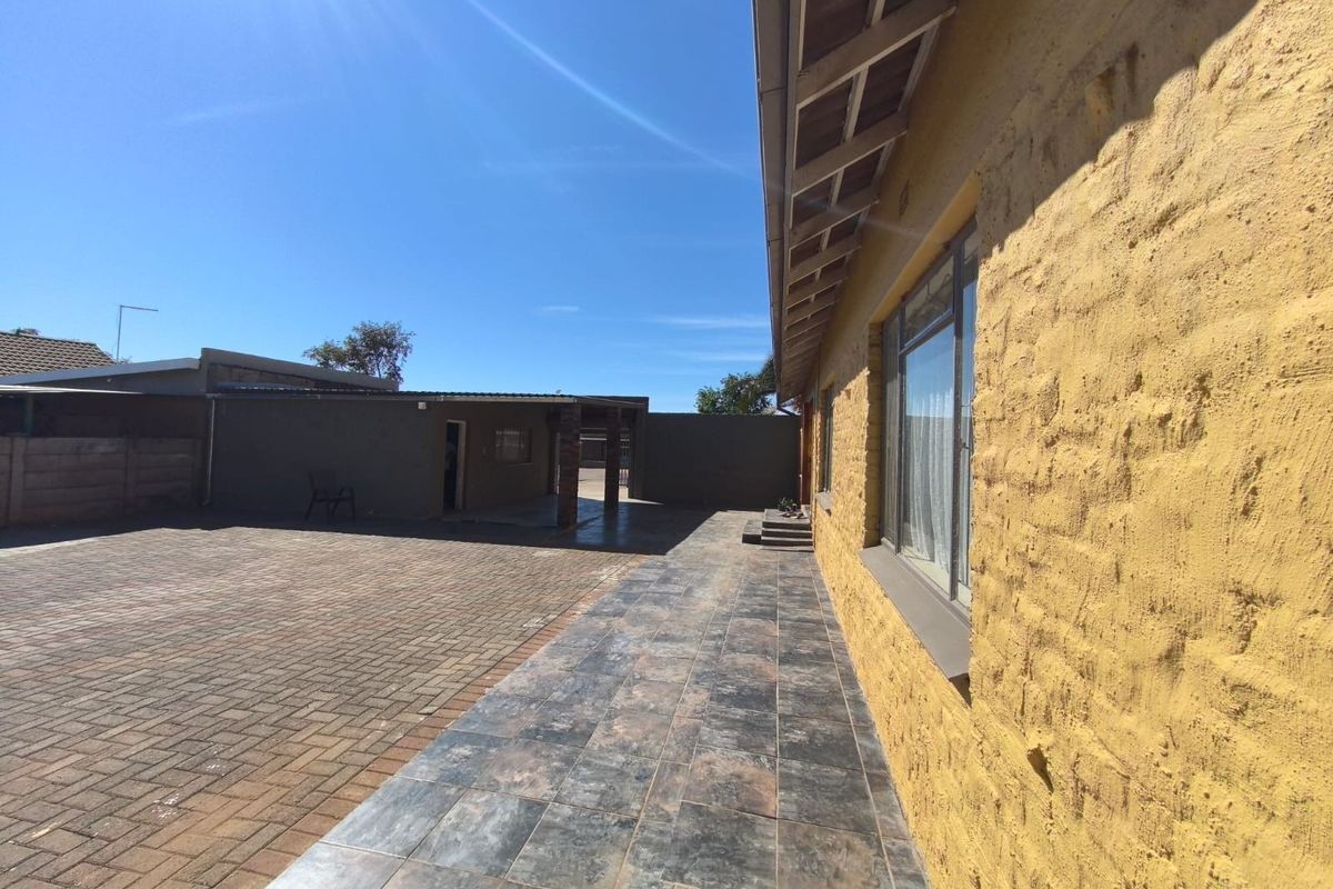 A rear exterior view featuring a paved driveway, a tiled patio area, and a textured yellow brick wall with exposed rafter eaves.