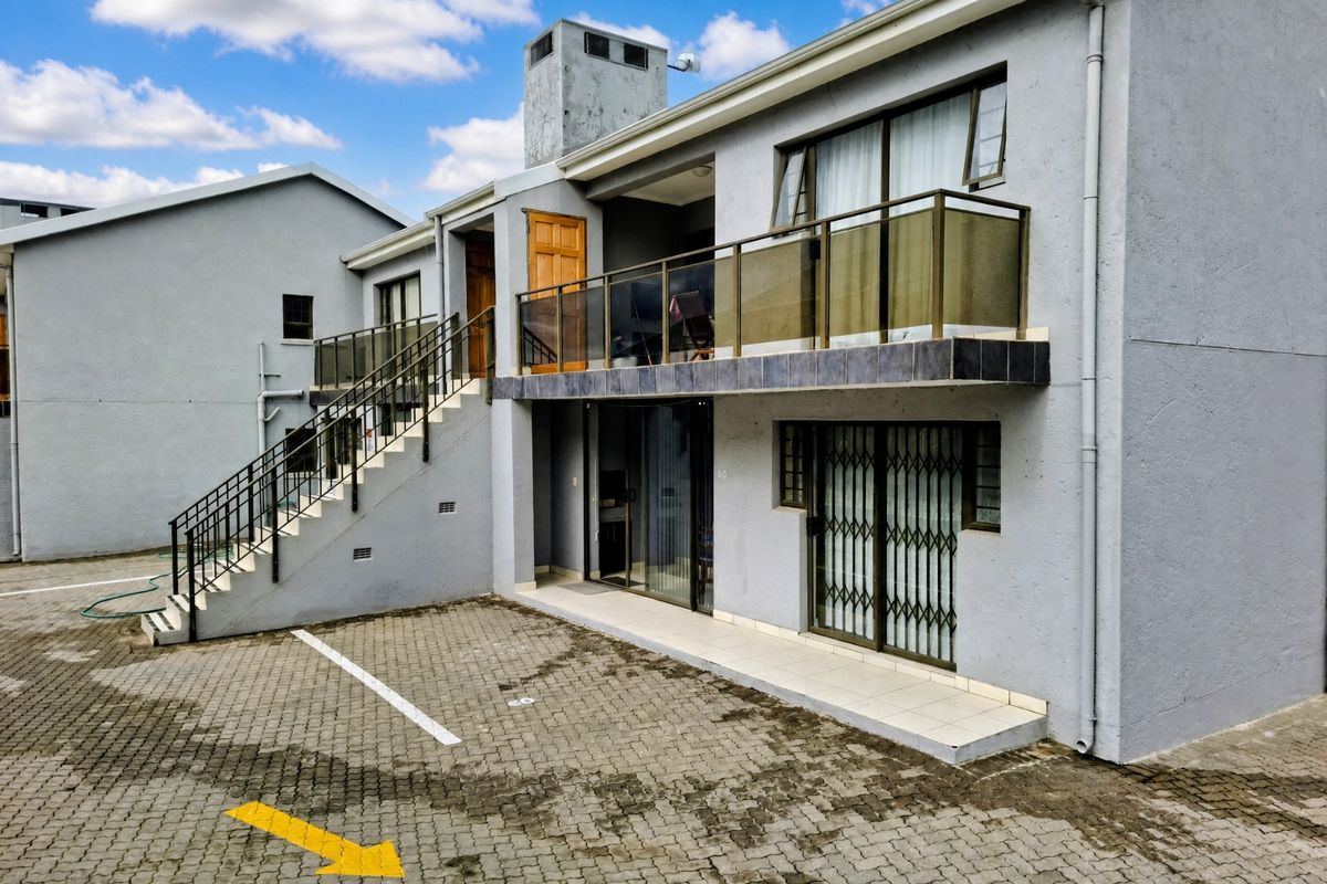 Exterior view of an apartment building featuring a paved parking area, external staircase, and a balcony with glass balustrades.