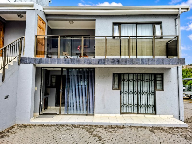 Front elevation of a two-story apartment featuring a balcony with glass balustrade, sliding doors, security gates, and a paved driveway.