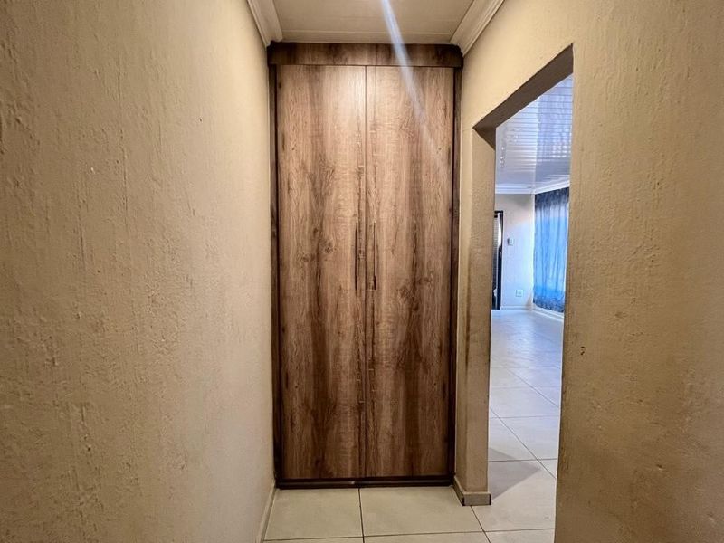 A narrow hallway featuring a built-in wooden cupboard and tiled flooring, leading into an adjacent living space.