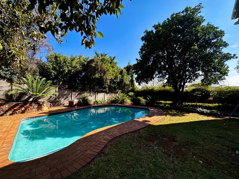 A private backyard featuring a swimming pool with brick coping, surrounded by established trees, garden beds, and a timber boundary fence.