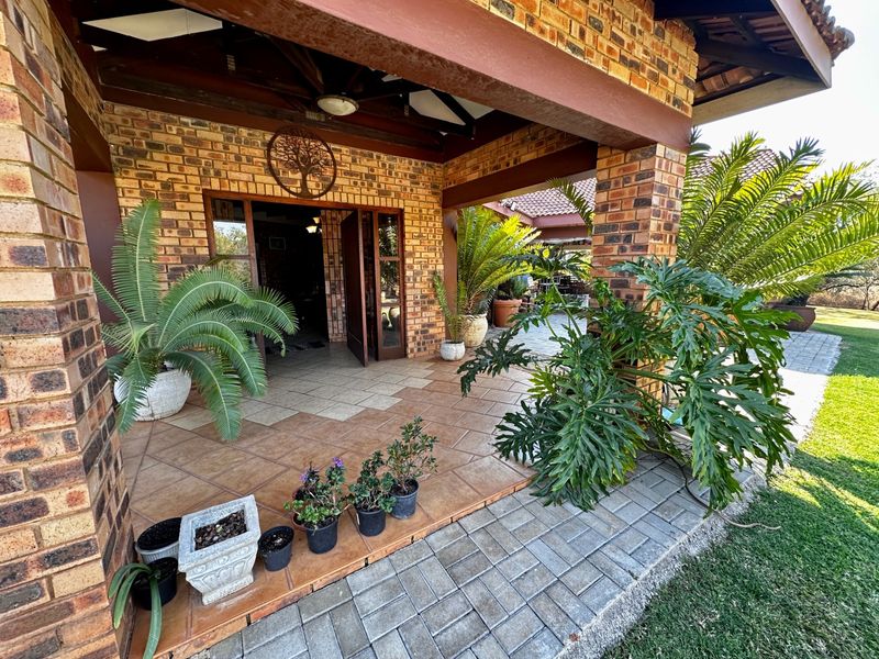 A covered patio area featuring exposed brick pillars, tiled flooring, and timber rafters, providing a seamless transition to the surrounding garden and paved walkway.
