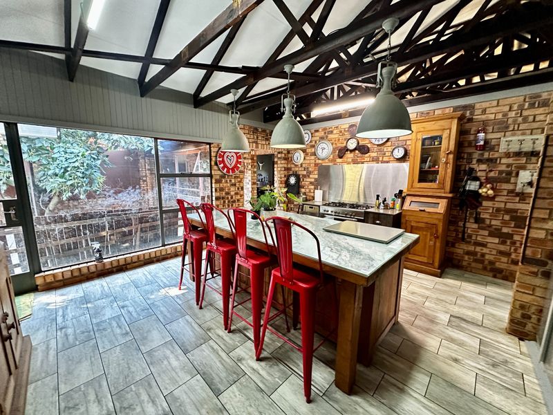 Rustic kitchen featuring exposed brick walls, timber roof trusses, a large central island with stone countertop, and modern tiled flooring.