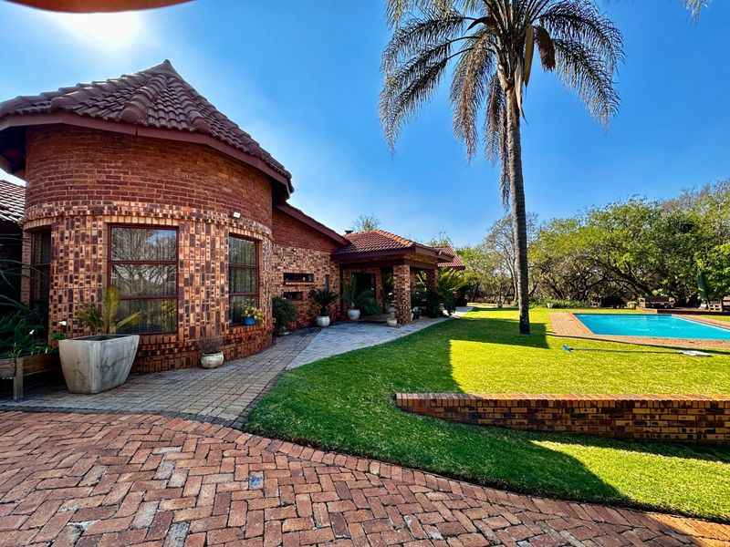 Rear view of a brick farmhouse featuring a swimming pool, manicured lawn, and a covered patio area set within a landscaped garden.