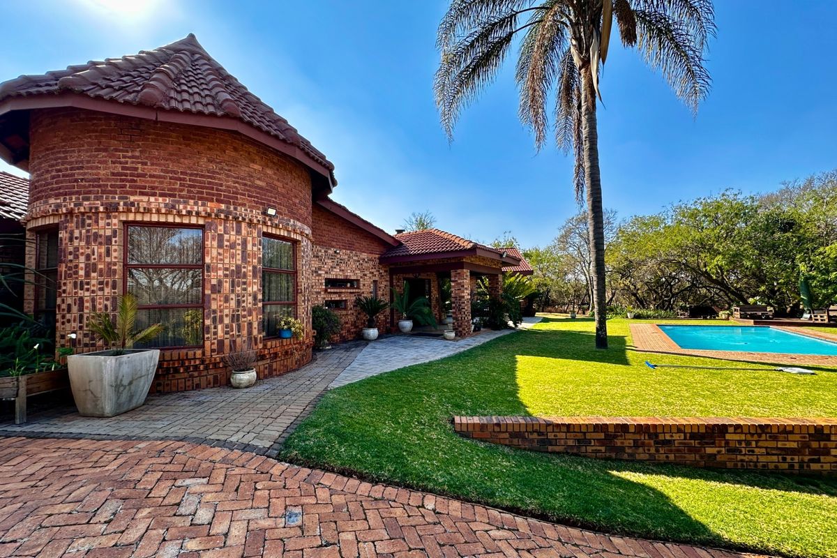 Rear view of a brick farmhouse featuring a swimming pool, manicured lawn, and a covered patio area set within a landscaped garden.