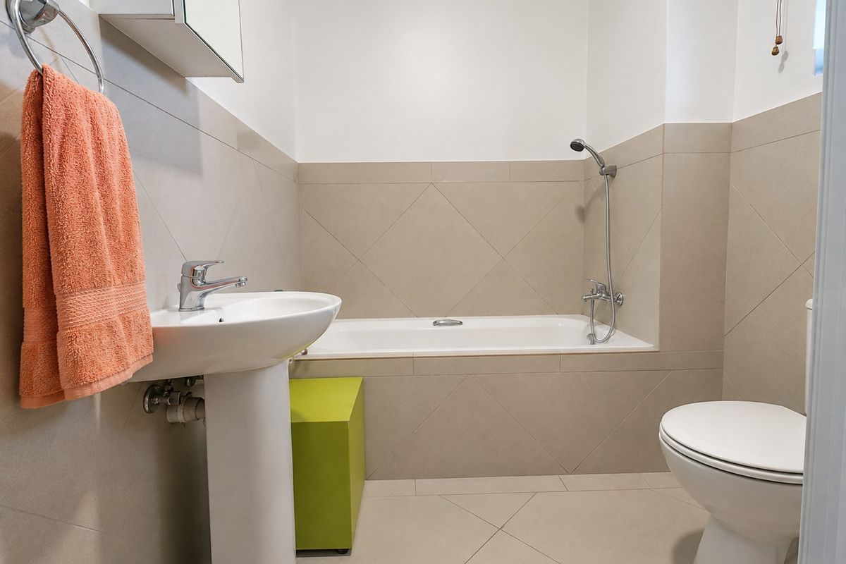 A clean, functional bathroom featuring a pedestal washbasin, a bathtub with a handheld shower attachment, and a standard toilet with neutral wall tiling.