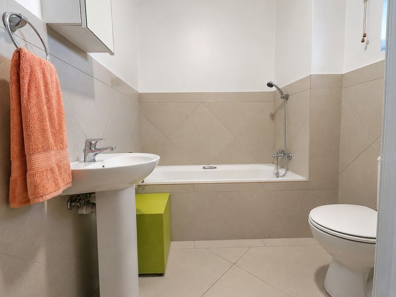 A clean, functional bathroom featuring a pedestal washbasin, a bathtub with a handheld shower attachment, and a standard toilet with neutral wall tiling.