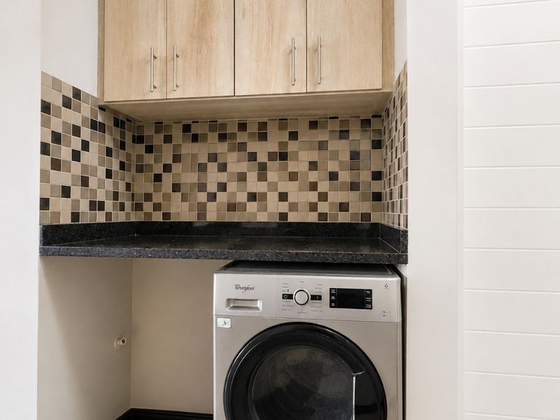 A compact, well-maintained laundry nook featuring built-in light wood cabinetry, a dark granite countertop, and a mosaic tile backsplash with tiled flooring.